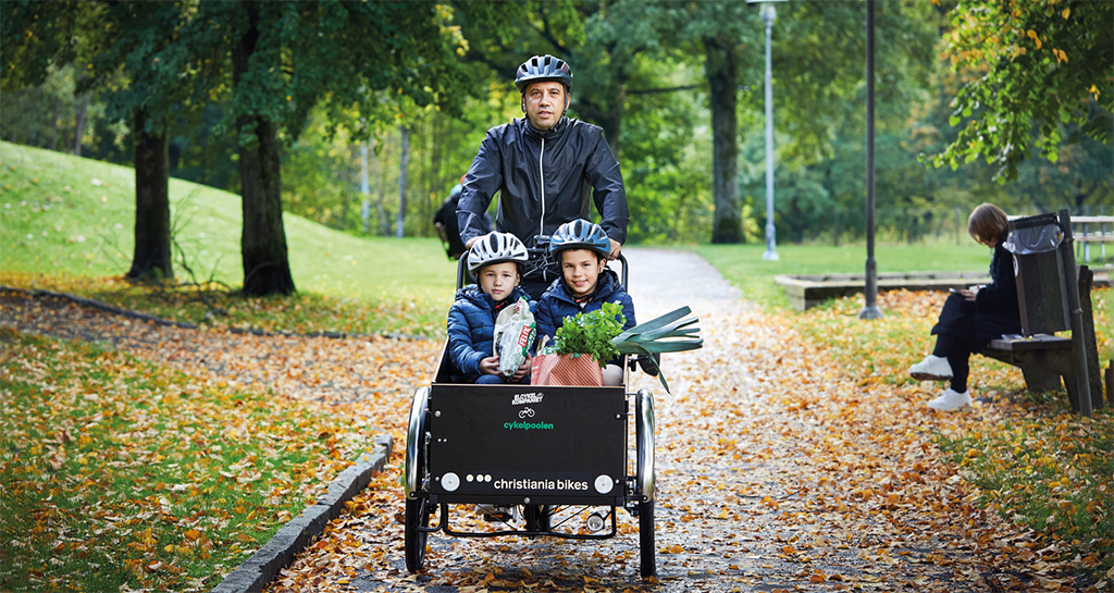 En man med två barn i en elpackcykel cyklar på en härlig cykelväg bland tröt, gräs och löv. Någon sitter på en parkbänk bakom.