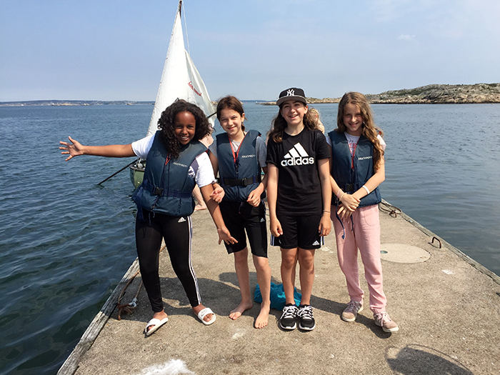 Fyra glada tjejer på en brygga med hav och en segelbåt i bakgrunden